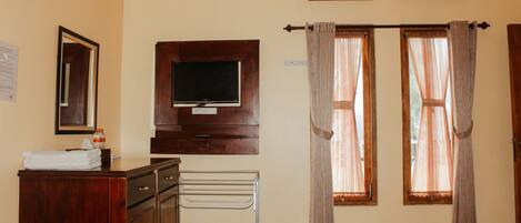 A hotel room with a wooden dresser, a TV mounted on the wall, a towel rack, and two windows with curtains.