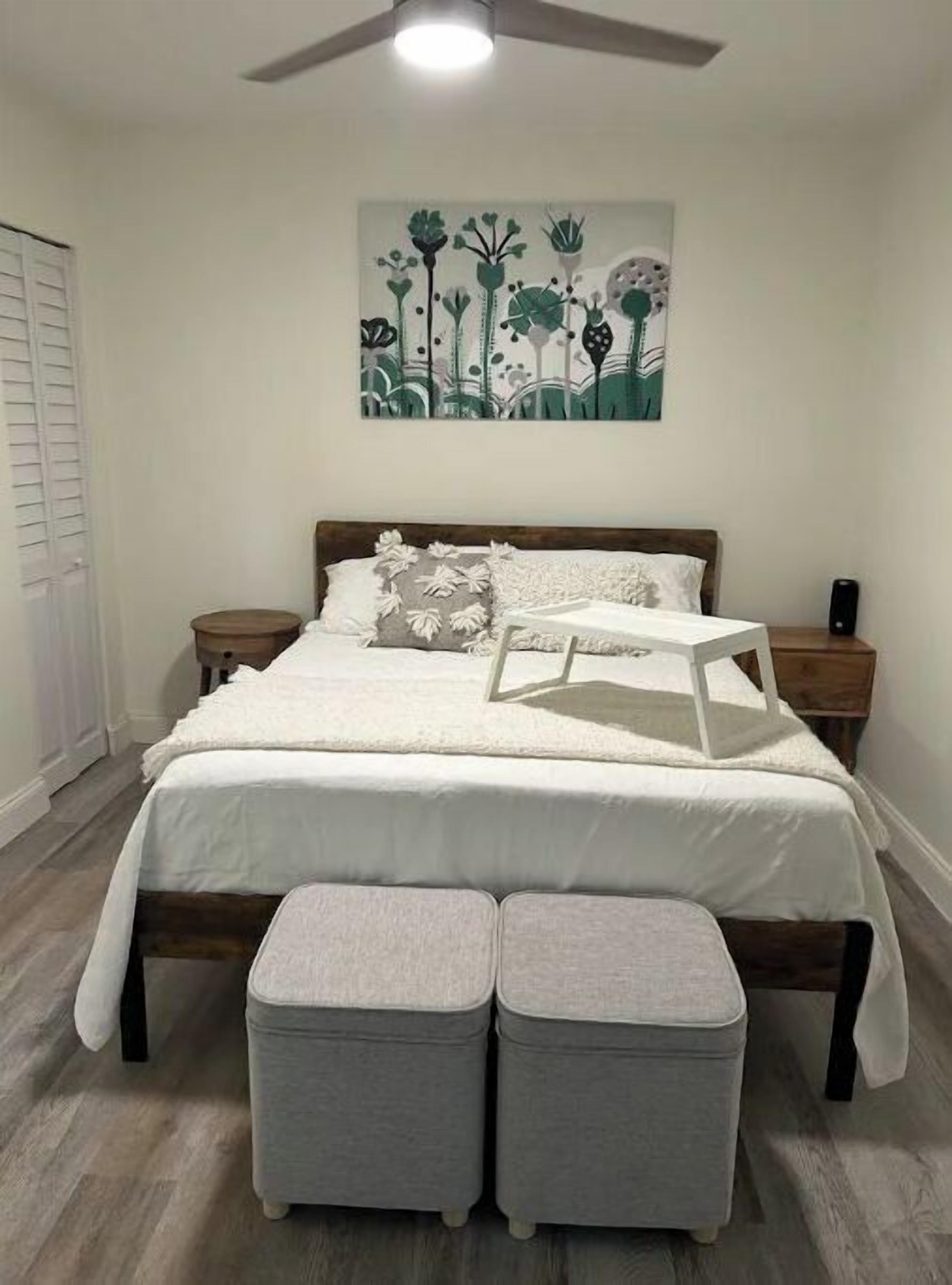 Ein Schlafzimmer mit einem Bett, zwei Nachttischen, einem Deckenventilator, einem Wandbild und einem Fenster mit Jalousien.