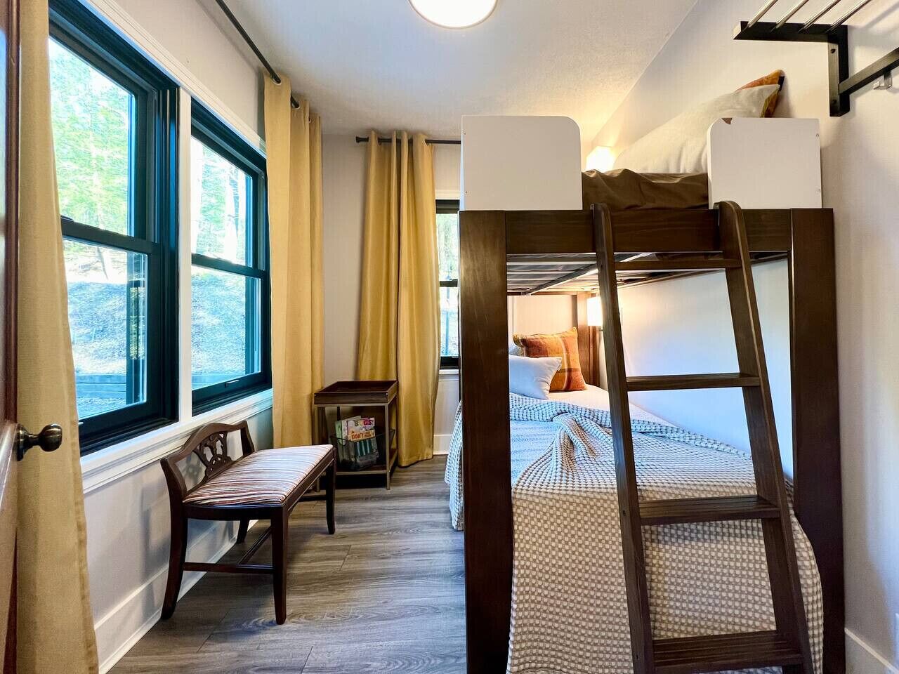A narrow room with a bunk bed, a small table, and a chair. There is a window on one side and a door on the other.