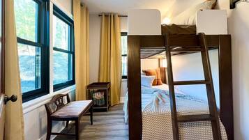 A narrow room with a bunk bed, a small table, and a chair. There is a window on one side and a door on the other.