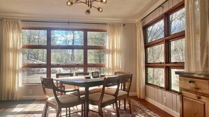 A dining room with a wooden table and chairs, large windows with curtains, and a chandelier.