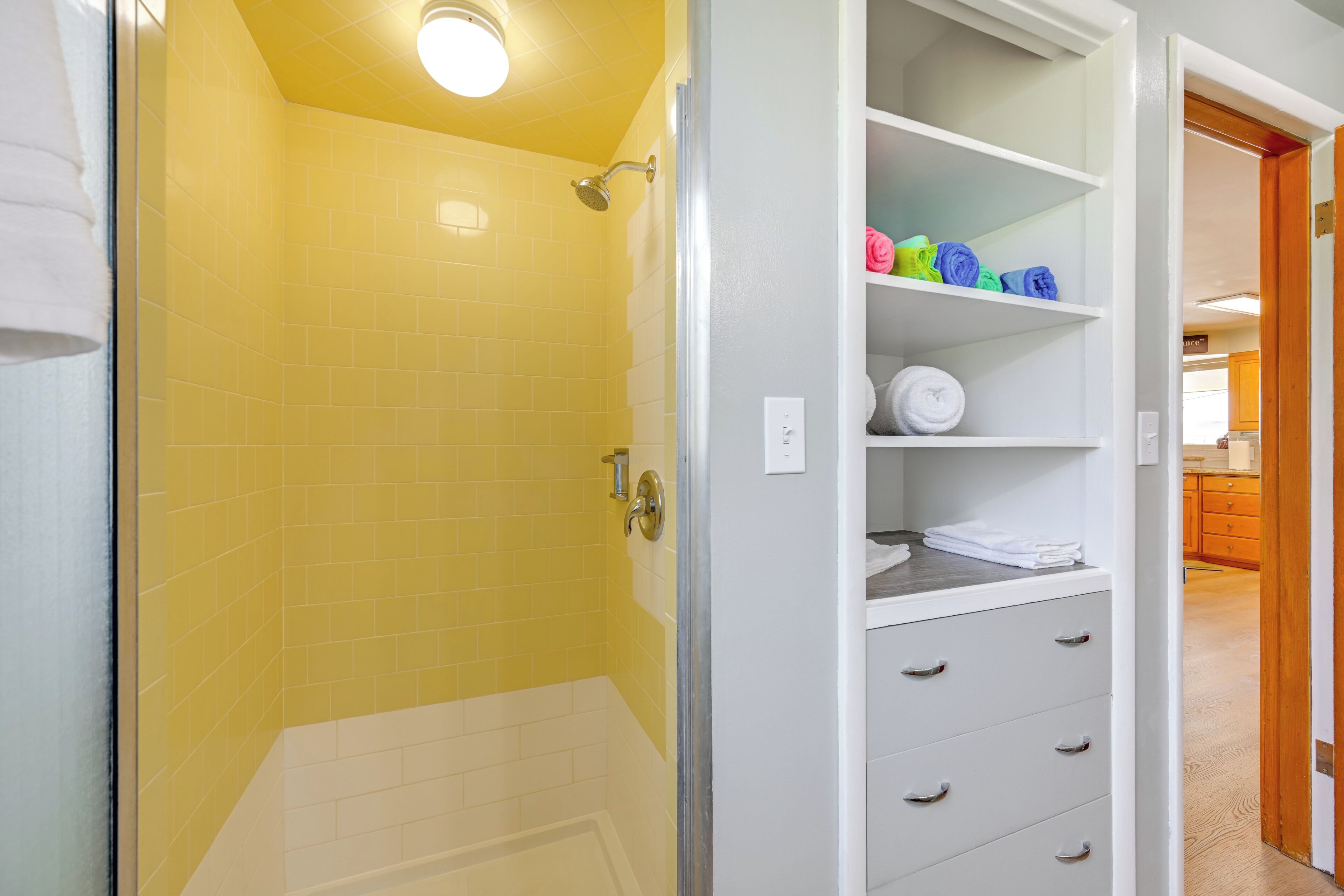 Un baño con ducha de azulejos amarillos, bañera blanca y un mueble de almacenamiento con estantes y cajones.