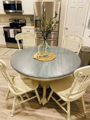Una mesa de comedor con un florero, rodeada de sillas blancas, en una cocina con electrodomésticos de acero inoxidable.