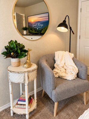 Ein gemütliches Zimmer mit einem grauen Sessel, einem kleinen runden Tisch mit Pflanze und Dekoration, einem Wandspiegel und einem Fernseher, der eine Sonnenuntergangsszene zeigt.