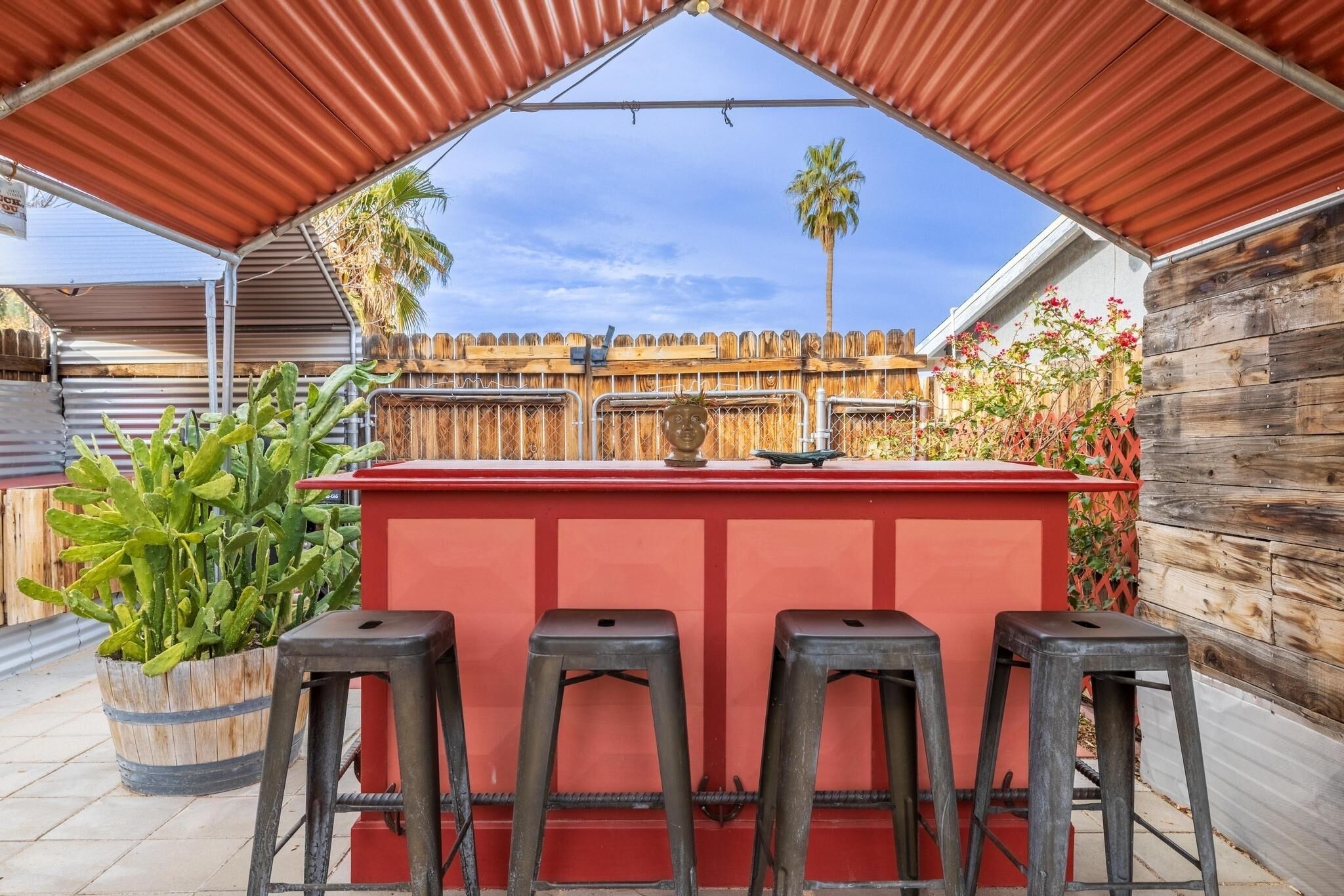 Un espace bar extérieur doté d’un comptoir rouge et de quatre tabourets, entouré d’une clôture en bois et de plantes.