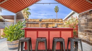 Zona de bar al aire libre con mostrador rojo y cuatro taburetes, rodeada por una valla de madera y plantas.