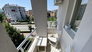 Un balcón con sillas blancas y una mesita, con vista a una calle con coches aparcados y edificios.