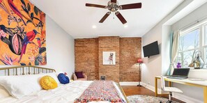 Ein Schlafzimmer mit Backsteinwand, einem Bett mit bunten Kissen, einem Schreibtisch mit Laptop und einem großen Fenster.