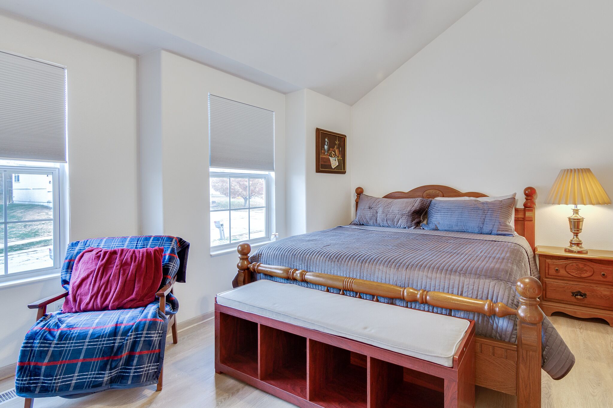 A bedroom with a bed, a chair with a blanket, a bedside table with a lamp, and a window with blinds.