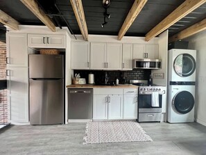 A modern kitchen with white cabinets, stainless steel appliances, a brick wall, and exposed wooden beams.