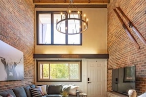 A loft-style room with exposed brick walls, a large window, a chandelier, a flat-screen TV, and a fireplace.