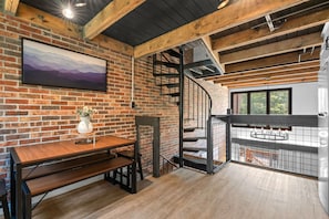 Un interior rústico con paredes de ladrillo visto, una mesa de comedor de madera, una escalera y una vista por una ventana.
