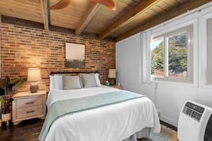 Un dormitorio con pared de ladrillo, cama con ropa blanca, mesita de noche, lámpara, ventana con vista a follaje y calentador blanco.