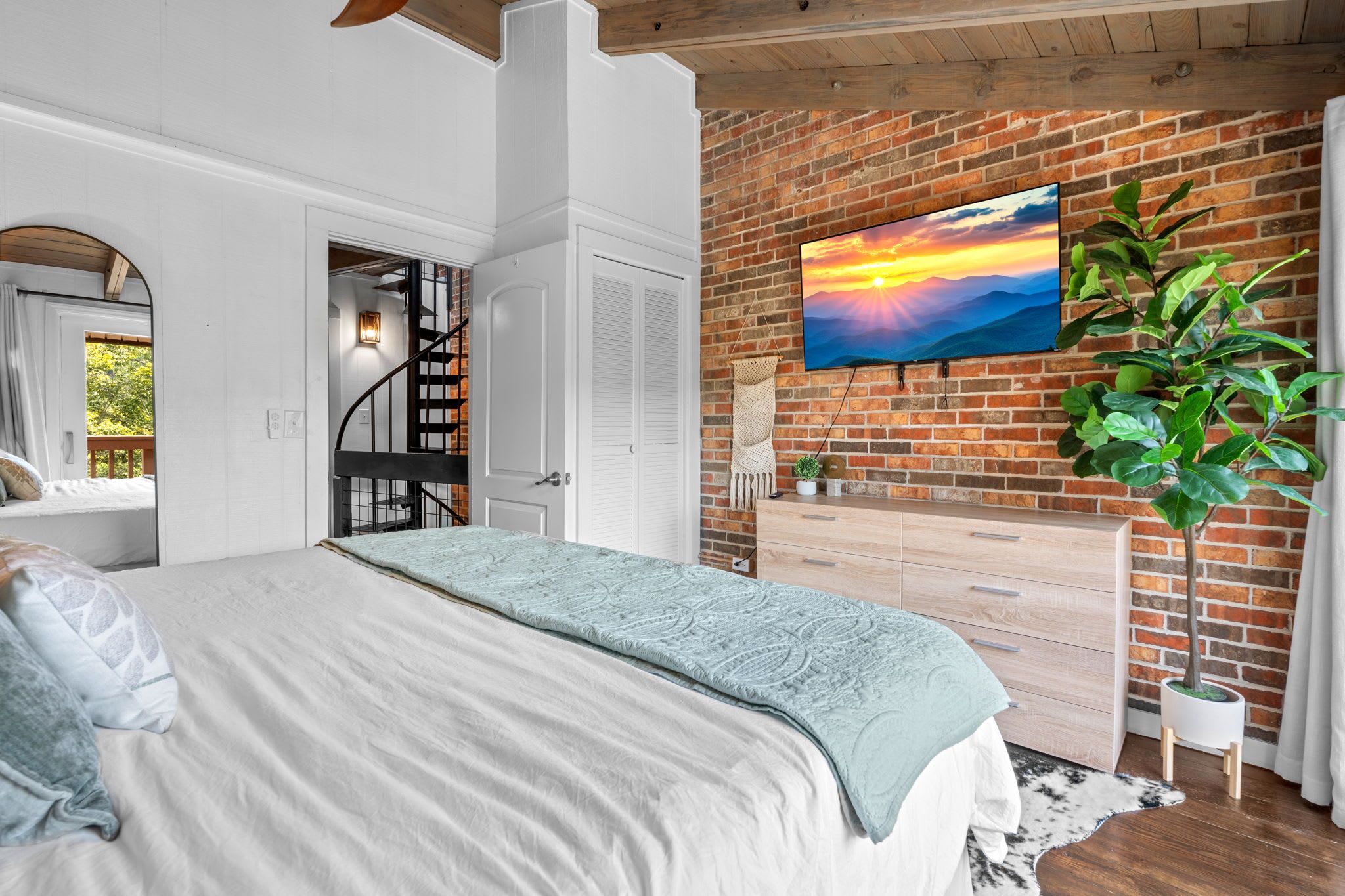 Un dormitorio con una cama, una escalera, un televisor montado en una pared de ladrillo y una planta en maceta.
