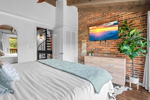 Une chambre avec un lit, un escalier, un téléviseur fixé au mur en briques et une plante en pot.