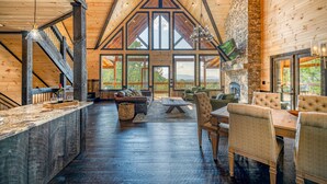 A spacious interior with a high ceiling, wooden beams, a stone fireplace, and large windows offering views of the outdoors. - Blue Ridge Luxury Cabin with Sunset Views (Mineral Bluff)