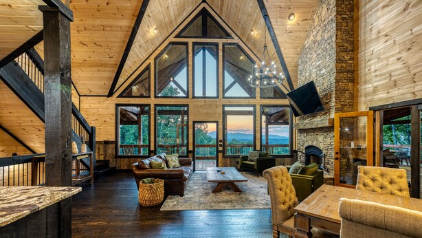 A spacious loft-style room with a high ceiling, large windows, and a stone fireplace. - Blue Ridge Luxury Cabin with Sunset Views (Mineral Bluff)
