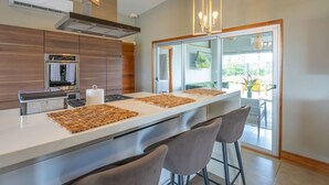 A modern kitchen with a central island, bar stools, a built-in oven, and a large sliding glass door leading to an outdoor area. - Breathtaking Ocean View Villa, Monkeys Daily, Private Gated Development (Manuel Antonio)