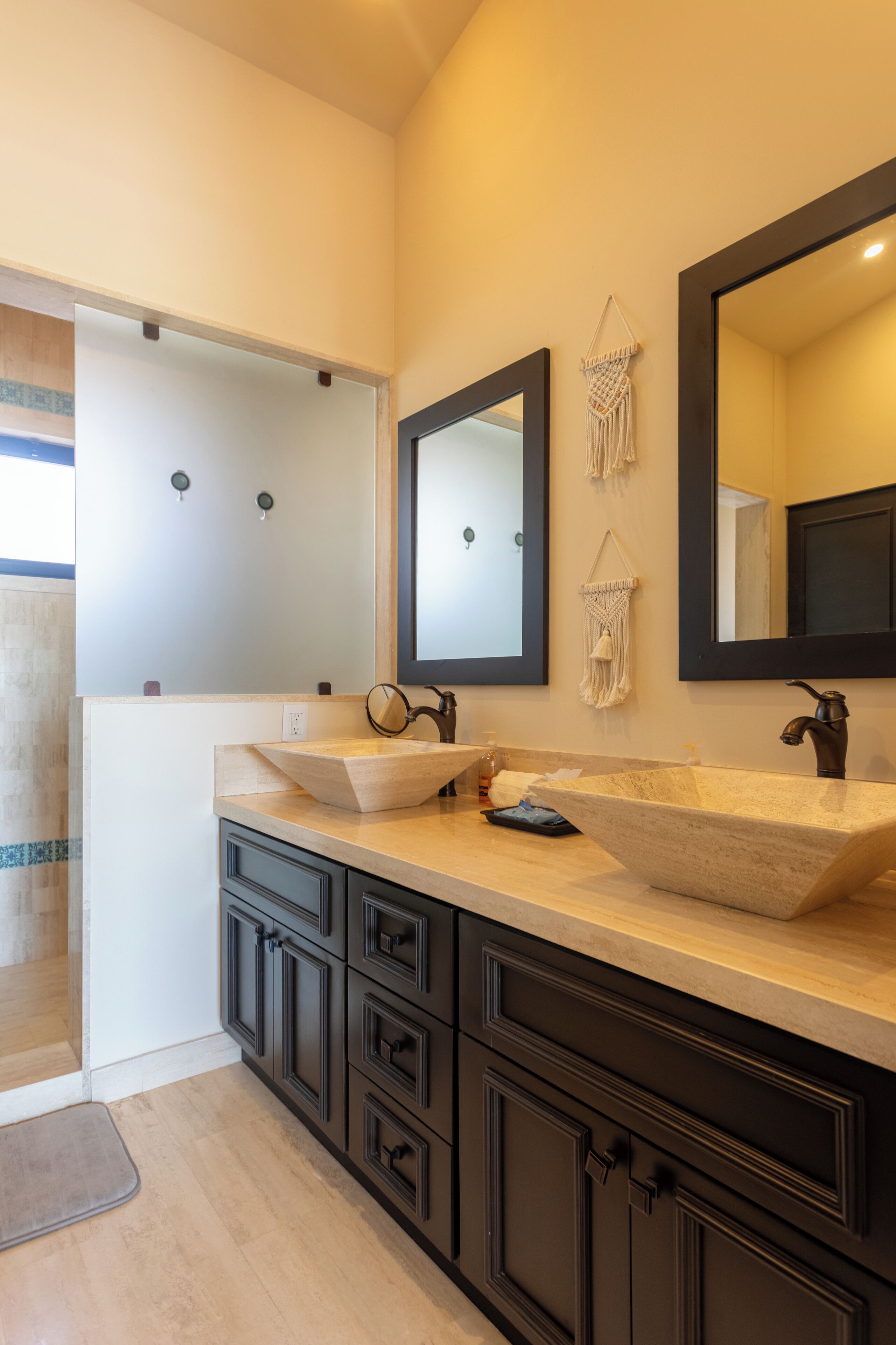 A bathroom with two sinks, dark cabinets, a large mirror, and a macrame wall hanging.