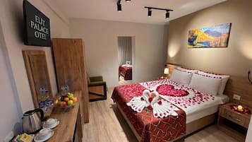 A hotel room with a bed adorned with rose petals and a swan decoration, a wooden side table with a fruit bowl, a kettle, and cups, and a painting on the wall.