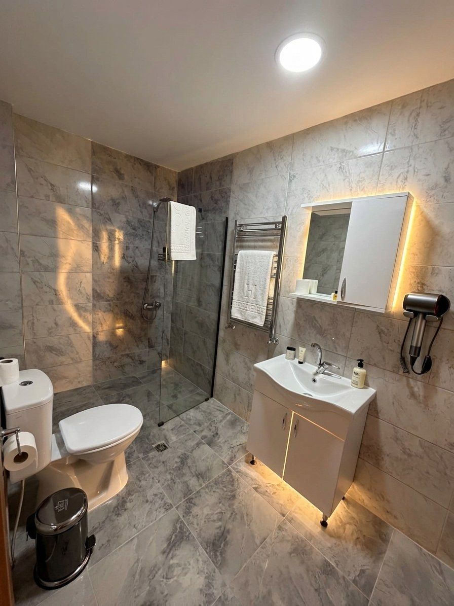 A modern bathroom with a glass-enclosed shower, a white sink with a mirror above, and a toilet with a paper towel holder.