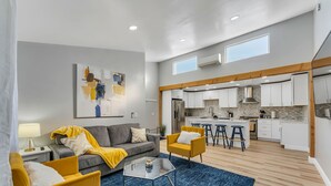 A modern living room with a sofa, armchairs, and a coffee table. A kitchen with white cabinets and a stone backsplash is visible in the background. - Open calender. Washer/dryer. Complementary Netflix. Struck Gold (Reno)