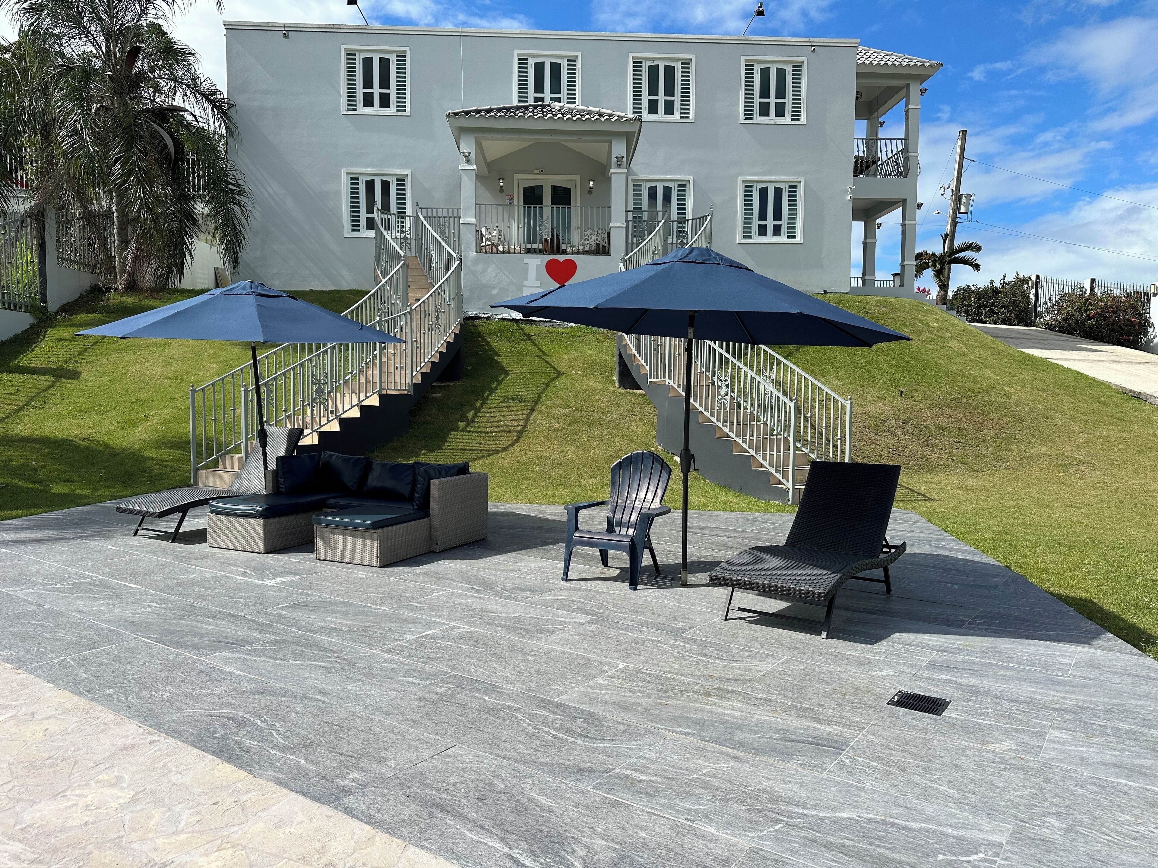 A two-story building with a staircase, outdoor seating area with chairs and a sofa, and a large umbrella providing shade.