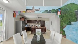 Un comedor con una mesa de vidrio y sillas blancas, una cocina al fondo, y un mural con una escena costera en la pared.