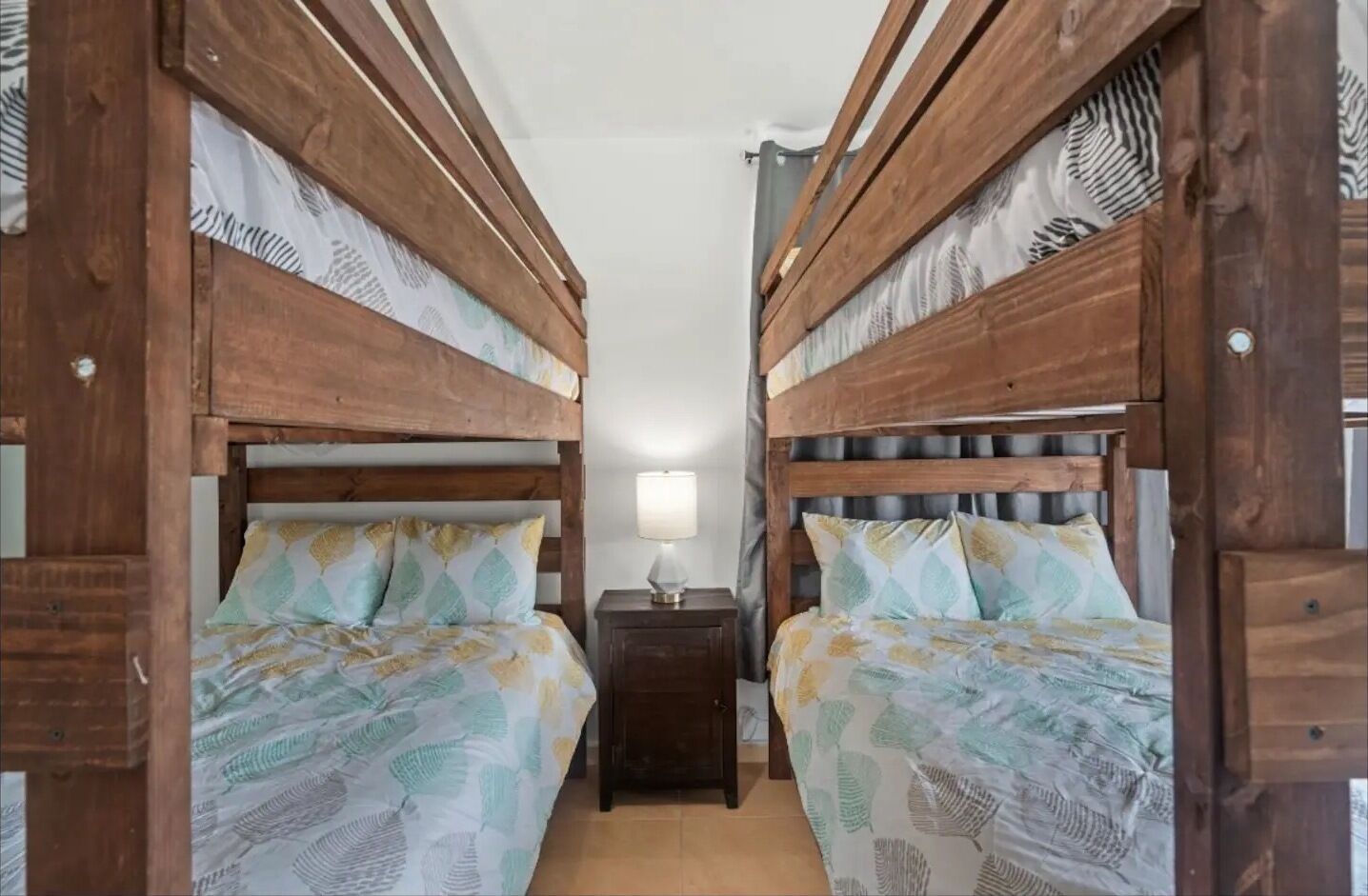 Une chambre avec deux lits superposés en bois, chacun agrémenté de draps à motifs de feuilles. Une petite table de chevet avec une lampe se trouve entre les lits.