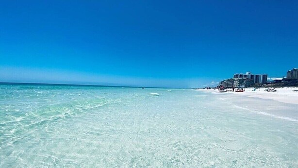 En strand med kristallklart turkost vatten, en sandstrand och en rad byggnader i bakgrunden.