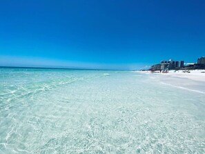A beach with clear turquoise water, a sandy shore, and a row of buildings in the background.