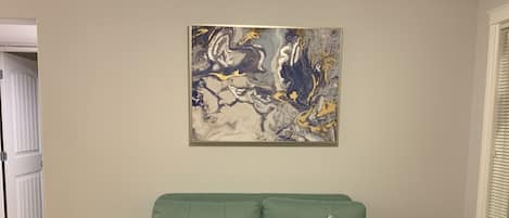 A green sofa with two patterned pillows, a wooden side table with two remotes, and a large abstract painting on the wall.