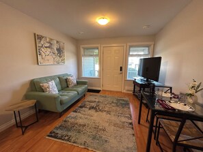 A living room with a green sofa, a flat-screen TV, a dining table with chairs, and a large abstract painting on the wall.