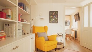 Ein leuchtend gelber Sessel mit einem grauen Kissen, ein weißes Bücherregal mit Büchern und Dekorationsobjekten, ein kleiner Tisch mit einem Korb und ein an der Wand befestigtes Bild von Blumen.