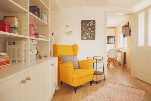 A bright yellow armchair with a grey pillow, a white bookshelf with books and decorative items, a small table with a basket, and a wall-mounted picture of flowers.