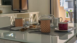 Una mesa de comedor con tazas de café, un plato de pasteles y una jarra, en una habitación con televisión y vistas al exterior.
