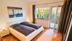 Een slaapkamer met een bed, nachtkastje en een raam met uitzicht op bomen en een stadsbeeld.