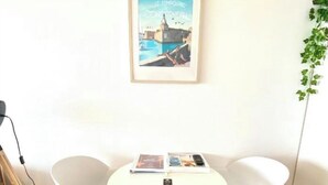 Ein Zimmer mit einem runden weißen Tisch, zwei weißen Stühlen, einem gerahmten Bild an der Wand und einer Pflanze rechts.