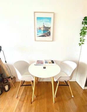 A room with a round white table, two white chairs, a framed picture on the wall, and a plant on the right.