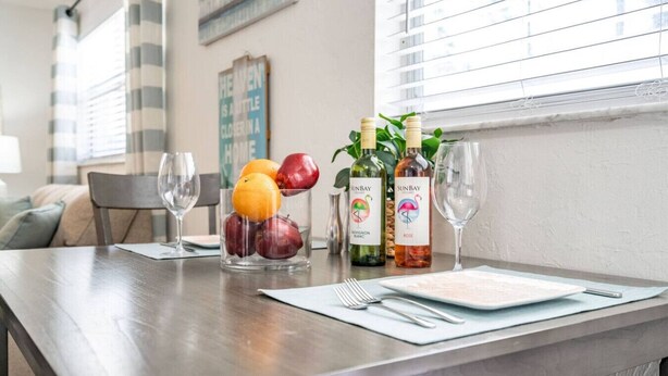 Uma mesa de jantar com uma tigela de frutas, garrafas de vinho e copos.