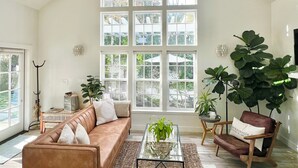 A spacious living room with a high ceiling, large windows, a brown leather sofa, a glass coffee table, and a large plant. - Spa Like Stay at the Award Winning Britt Bungalow in the Heart of Jacksonville (Jacksonville)