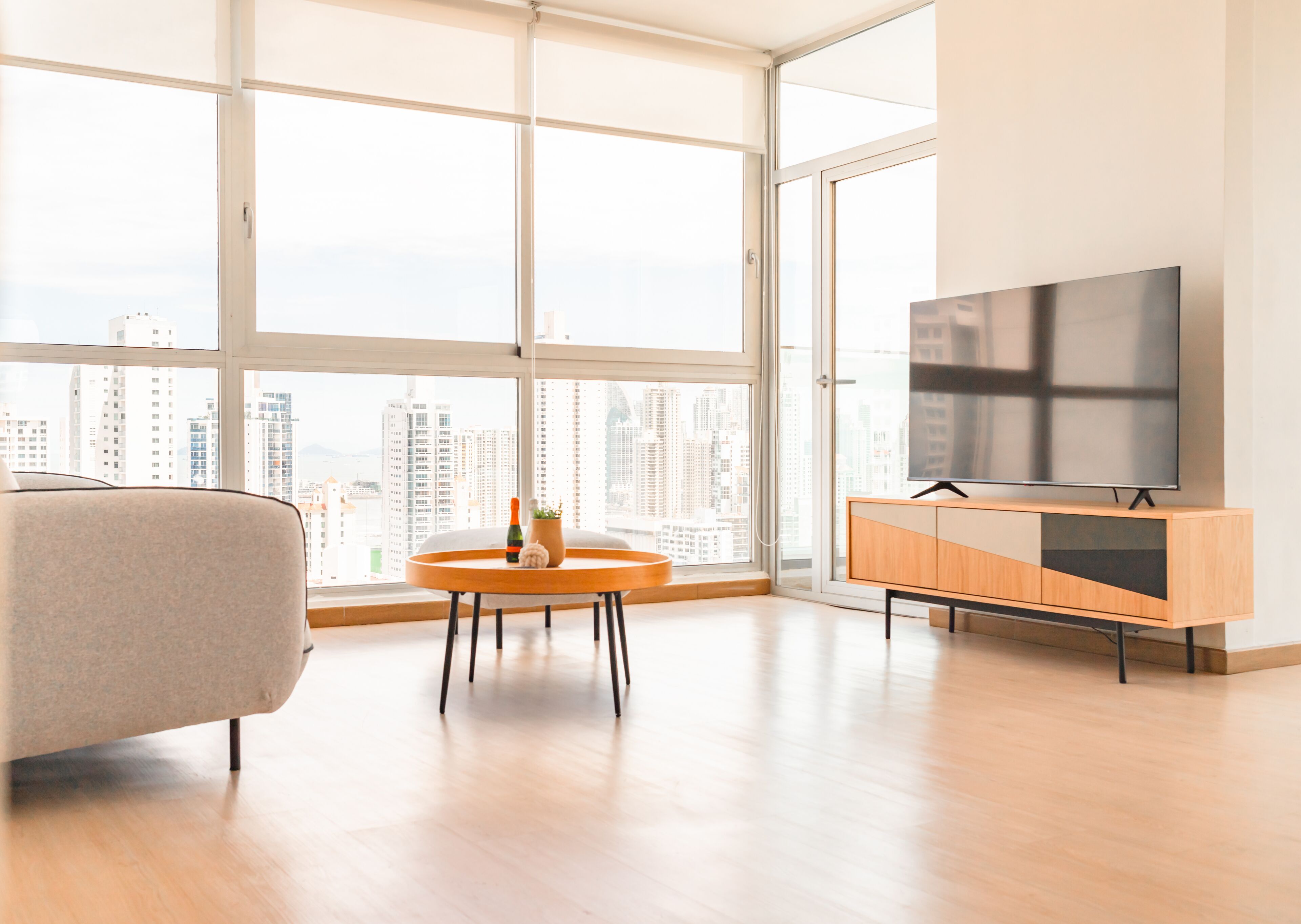 Una sala de estar moderna con un ventanal grande, un sofá, una mesita redonda con una botella y un objeto decorativo, y un televisor sobre un mueble de madera.