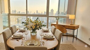 A dining area with a table set for four, large windows with a cityscape view, a small side table with a lamp, and a television on a stand. - Boutique Apartments Panamá Coco del Mar (Panama City)