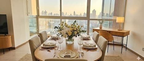 A dining area with a table set for four, large windows with a cityscape view, a small side table with a lamp, and a television on a stand.