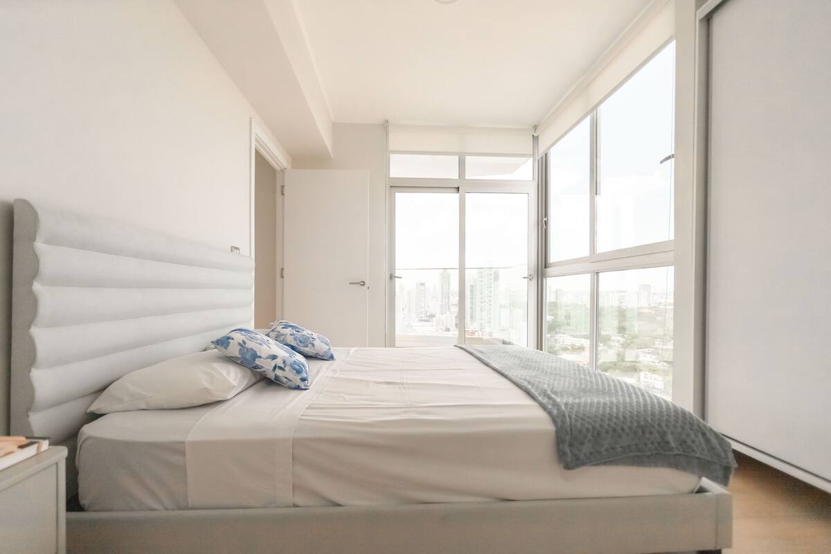 Ein Schlafzimmer mit großem Fenster, einem Bett mit weißer Bettwäsche und Blick auf eine Stadtlandschaft.