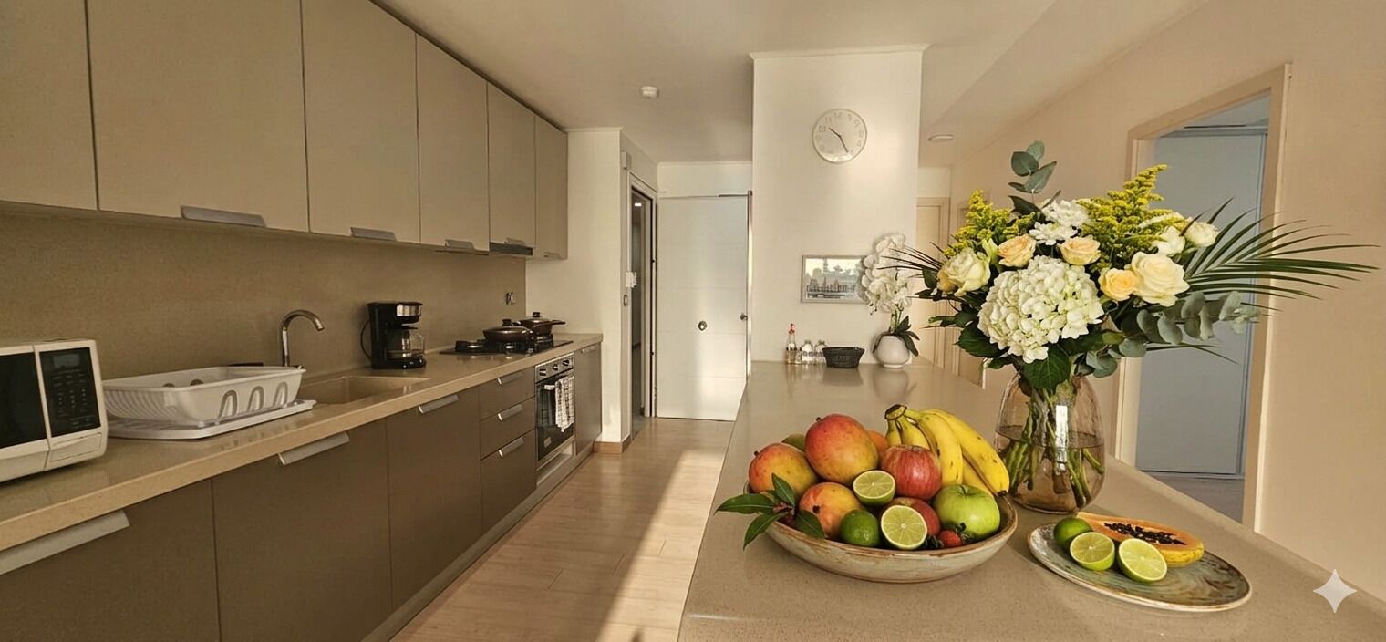 A modern kitchen with a microwave, stove, and sink. There is a fruit bowl with various fruits and a vase with flowers on the counter.
