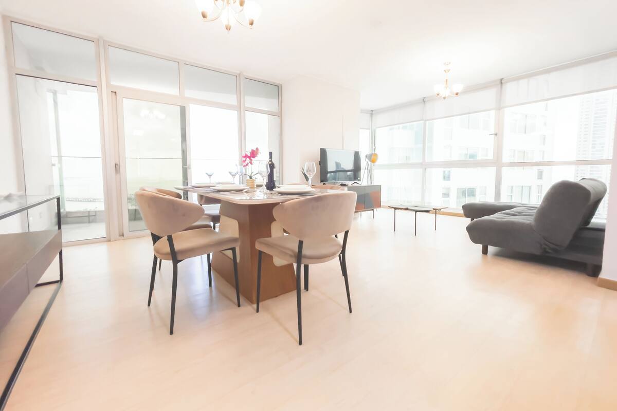 Ein modernes Wohnzimmer mit Essbereich, großen Fenstern und einem grauen Sofa.