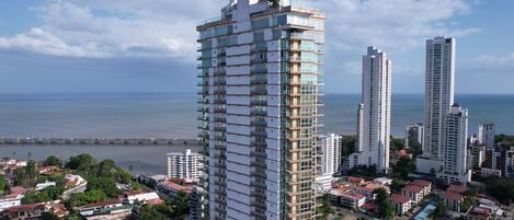A high-rise building with multiple balconies, surrounded by other buildings and a coastal view.