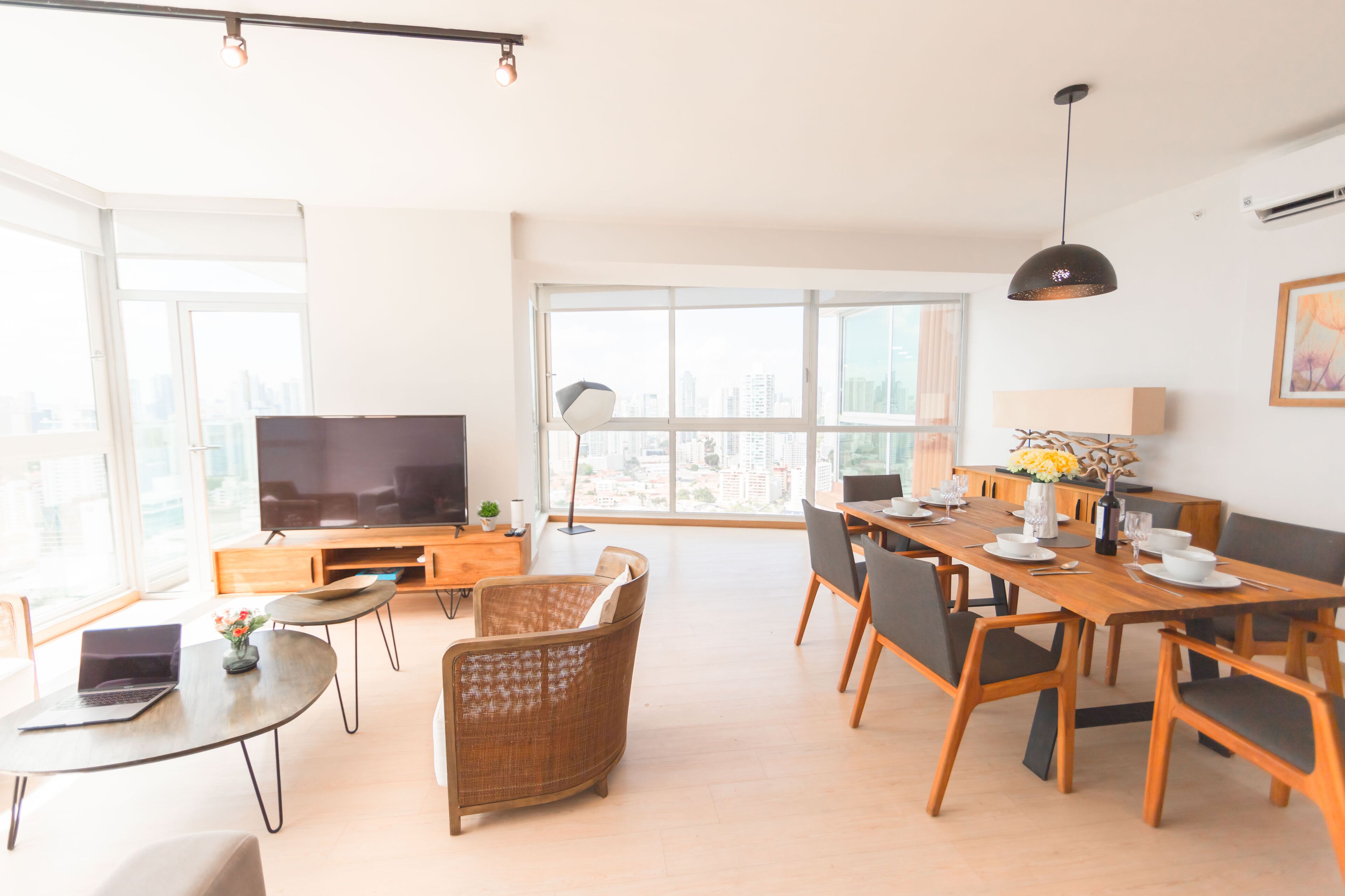 A modern living room with a large TV, dining table, and chairs, a balcony with a city view, and a large window.