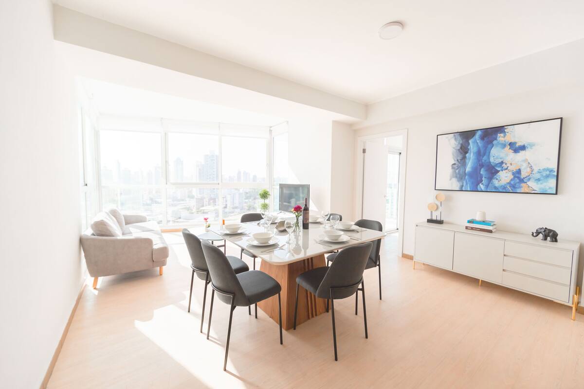 Ein moderner Essbereich mit einem großen Tisch, gedeckt für sechs Personen, umgeben von Stühlen. Es gibt eine Couch und eine Vitrine mit Dekorationsobjekten.